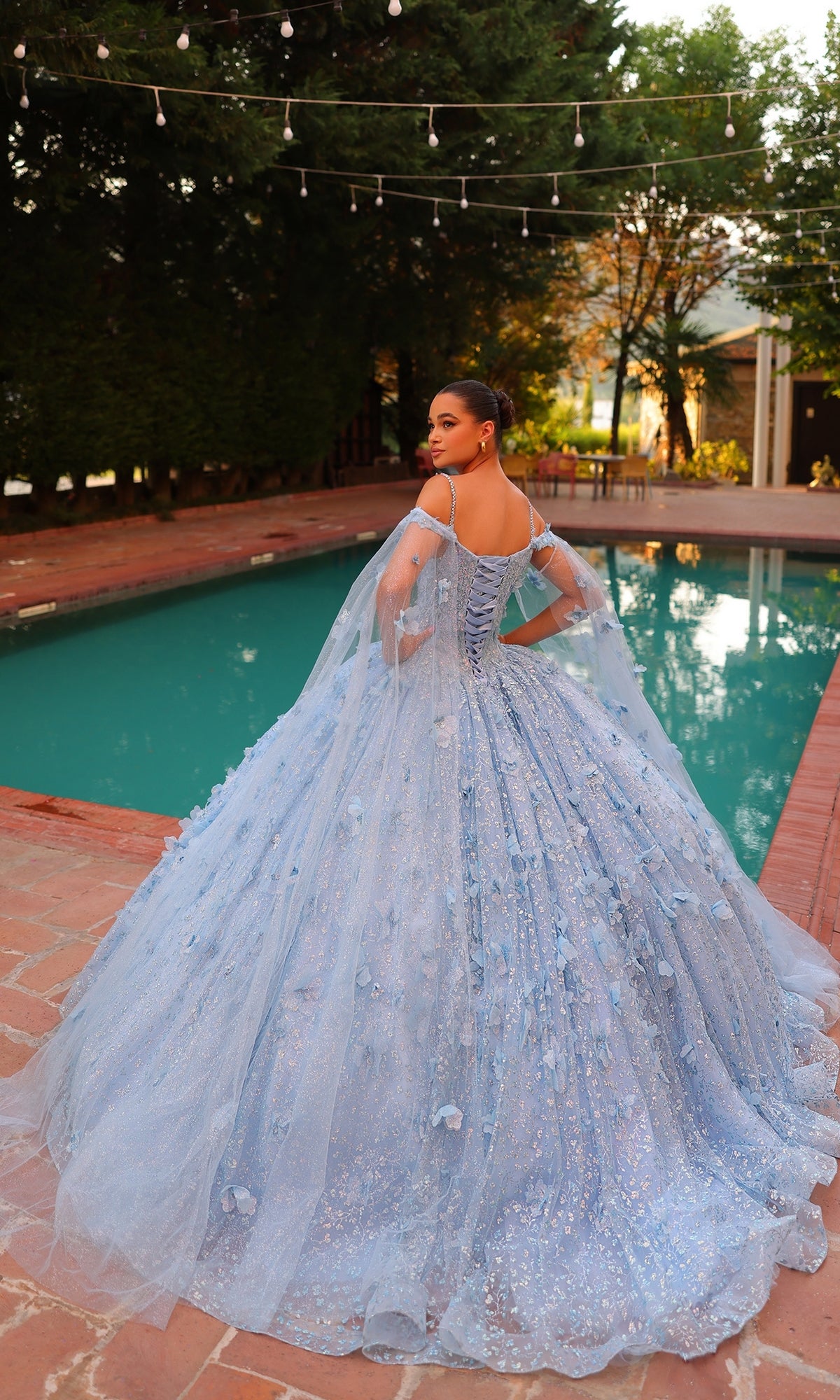 A woman in the Quinceanera Dress 54308 By Amarra?a blue, glittering off-the-shoulder ball gown with sheer cape sleeves?stands by a pool on a brick patio, facing away under string lights.